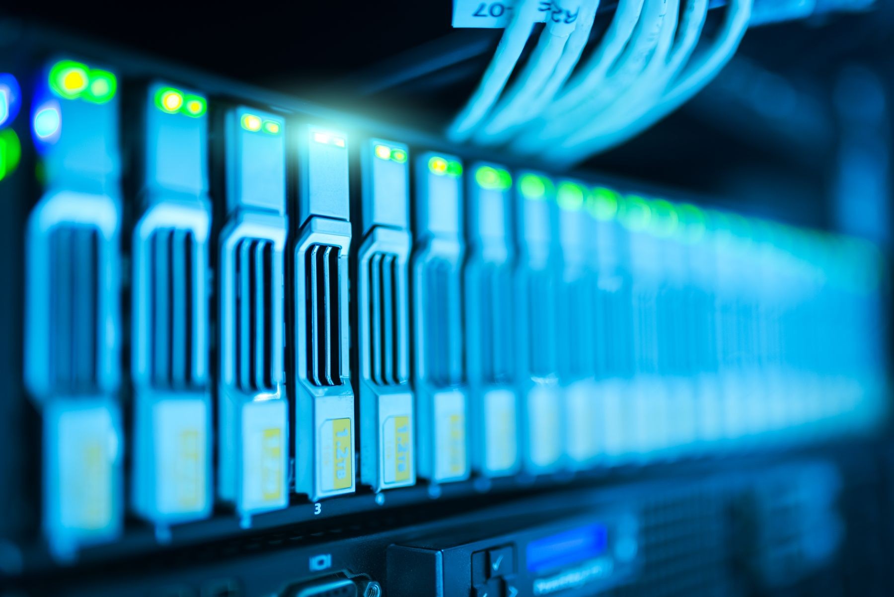 Primer plano de equipos en un rack, con indicadores verdes encendidos y cables claros en la parte superior, bajo iluminación azul.